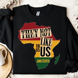 May include: Black sweatshirt with a map of Africa in red, yellow, and green. The text "THEY NOT LIKE US" is printed across the map. Below the map are the numbers "1865" and the word "JUNETEENTH".
