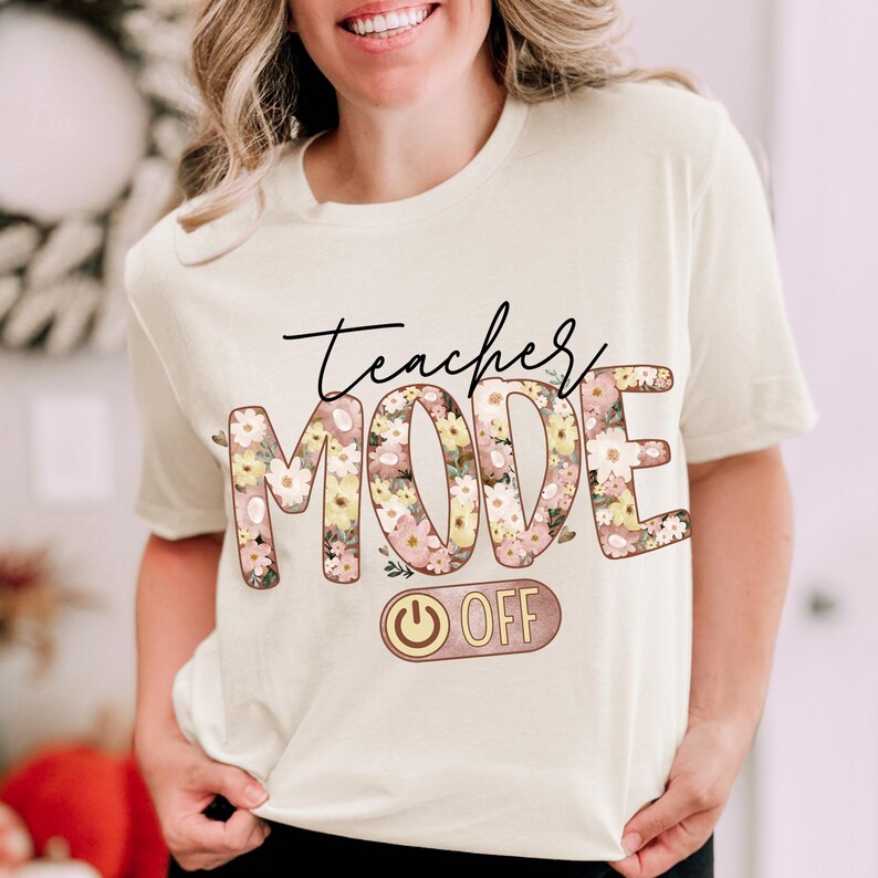May include: A white t-shirt with the text "teacher mode off" in pink and brown lettering. The word "mode" is decorated with pink and yellow floral designs. A pink and gold power button with the word "off" is below the text.