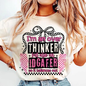 Puede incluir: Camiseta color crema con el texto "I'm an over THINKER but also an IDGAFER so it balances out." El texto es en negro y rosa, con un dise&ntilde;o de lazo a cuadros.