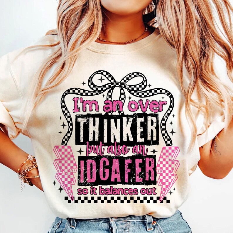 Puede incluir: Camiseta color crema con el texto "I'm an over THINKER but also an IDGAFER so it balances out." El texto es en negro y rosa, con un dise&ntilde;o de lazo a cuadros.