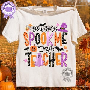 May include: A cream-colored short-sleeve t-shirt featuring the text "You can't SPOOK ME I'm a TEACHER" in colorful, patterned letters. The design includes a purple witch hat, a jack-o'-lantern, a ghost, bats, candy corn, and small stars.