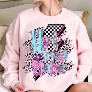 May include: Pink sweatshirt with a graphic design of the words "Hot Mess Mama" in a colorful, playful font with checkered, leopard print, and star accents.