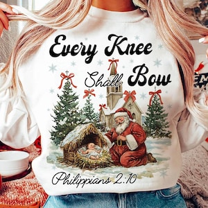 May include: White Christmas sweatshirt with a vintage illustration of a nativity scene. The text "Every Knee Shall Bow" is printed in black with a red bow above the word "knee". The text "Philippians 2:10" is printed below the illustration.