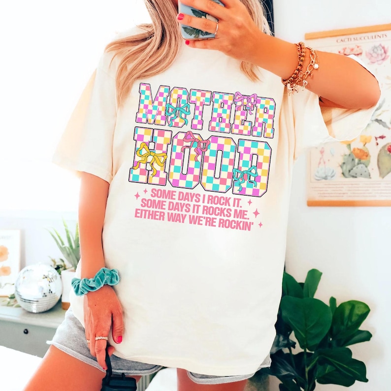 May include: A white t-shirt with a chequered design and the text "MOTHERHOOD" in pink, blue, and yellow. The text is surrounded by colourful bows. The text below the design reads "SOME DAYS I ROCK IT. SOME DAYS IT ROCKS ME. EITHER WAY WE'RE ROCKIN'."