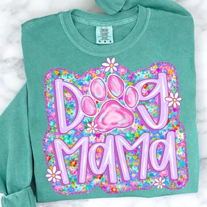 May include: A teal green t-shirt with a pink paw print and the text "Dog Mama" surrounded by colorful flowers.