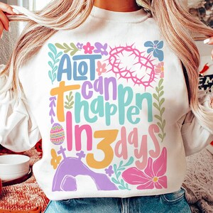 May include: White sweatshirt with a colorful graphic design that says "A lot can happen in 3 days". The graphic includes flowers, a crown of thorns, and a tomb.
