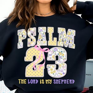 May include: Black sweatshirt featuring the text "PSALM 23" in a floral and plaid design. The number "23" is in a yellow polka dot pattern and floral design, with a pink ribbon. The text "THE LORD IS MY SHEPHERD" is below.