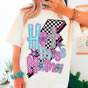 May include: White t-shirt with a graphic design that says "Hot Mess Mama" in pink, blue, and black letters with stars and checkered patterns.
