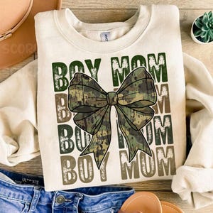 May include: A cream-colored sweatshirt with the text "BOY MOM" repeated four times in a distressed font. A large camouflage bow is centered between the words.