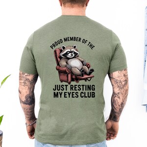 Op de afbeelding: Groen t-shirt met een cartoon wasbeer die in een stoel ontspant. De tekst luidt "Proud Member of the Just Resting My Eyes Club". De wasbeer is grijs, wit en zwart en de stoel is rood.