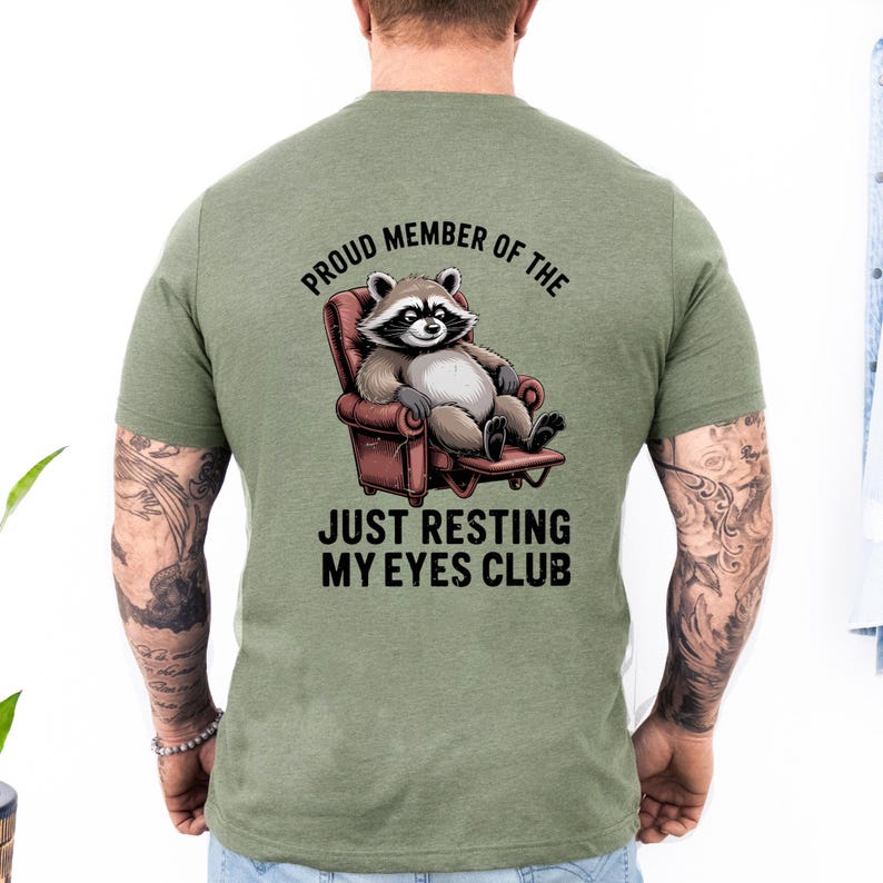 Op de afbeelding: Groen t-shirt met een cartoon wasbeer die in een stoel ontspant. De tekst luidt "Proud Member of the Just Resting My Eyes Club". De wasbeer is grijs, wit en zwart en de stoel is rood.