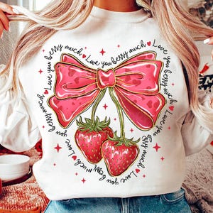 May include: White sweatshirt with a pink bow and two red strawberries. The text "Love you berry much" is repeated around the bow and strawberries.