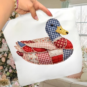 Peut inclure: Un coussin carré blanc avec un motif de canard en patchwork. Le canard a une tête à carreaux bleus et blancs, un bec jaune et un corps en tissu à motifs rouges, blancs et bleus. Le coussin est tenu par une main.
