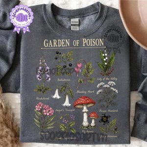 以下が含まれることがあります： 「GARDEN OF POISON」と題された植物画がプリントされたダークグレーのクルーネックトレーナー。トリカブト、ベラドンナ、ケマンソウ、スズラン、エンゼルトランペット、ドクニンジン、ベニテングタケ、クリスマスローズなどの植物のイラストが描かれており、それぞれに名前が記されています。