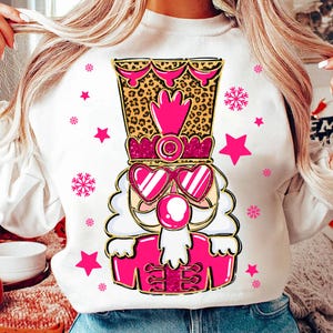 May include: White sweatshirt with a pink and gold nutcracker design. The nutcracker is wearing sunglasses and has a pink bubble coming out of its mouth. The design is surrounded by snowflakes and stars.