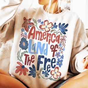 以下が含まれることがあります： 「America Land of the Free」というテキストを囲むように、赤、青、緑の花のグラフィックプリントが施された白いスウェットシャツです。