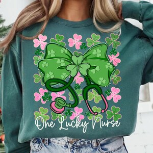May include: Teal green sweatshirt with a St. Patrick's Day theme. The design features a large green bow with a shamrock, a stethoscope, and pink and green shamrocks. The text "One Lucky Nurse" is written in white script.