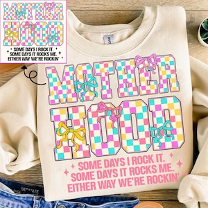 May include: A cream-coloured sweatshirt with a checkered design that spells out "MOTHERHOOD" in pink, blue, and yellow. The text "SOME DAYS I ROCK IT. SOME DAYS IT ROCKS ME. EITHER WAY WE'RE ROCKIN'" is printed below the design.