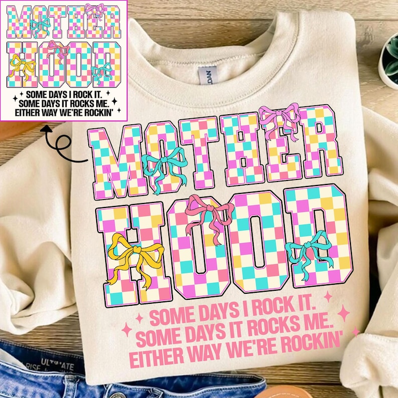 May include: A cream-coloured sweatshirt with a checkered design that spells out "MOTHERHOOD" in pink, blue, and yellow. The text "SOME DAYS I ROCK IT. SOME DAYS IT ROCKS ME. EITHER WAY WE'RE ROCKIN'" is printed below the design.