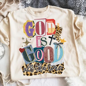 May include: A cream-coloured sweatshirt featuring the text "GOD IS GOOD all the time" in a collage of colourful letters and shapes. The design includes a cross, a heart, and a star, with a leopard print border.
