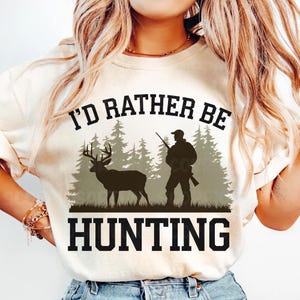 May include: A white t-shirt with the text "I'd rather be hunting" in black. The design features a silhouette of a hunter with a rifle and a deer in a forest setting.
