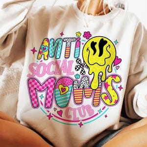 May include: Cream-colored sweatshirt with the text "ANTI SOCIAL MOMS CLUB" in a colorful, retro design. The design includes a melting yellow smiley face, hearts, stars, and checkered patterns. A fun, casual top.