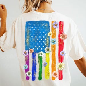 May include: White t-shirt with a graphic of an American flag with rainbow stripes and daisies.