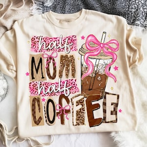 May include: Cream-colored sweatshirt with a graphic design. The design features the words "half MOM half COFFEE" in various fonts and patterns, along with a pink bow and a coffee drink illustration. Pink stars are scattered around the design.