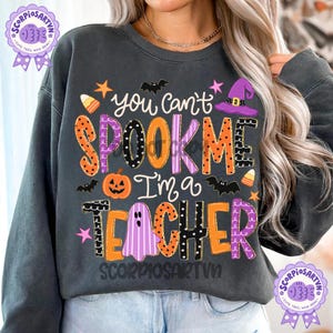 May include: A dark gray crewneck sweatshirt featuring the text "you can't SPOOK ME I'm a TEACHER" in colorful, patterned letters. The design includes Halloween-themed graphics such as a jack-o'-lantern, a purple witch hat, bats, and candy corn.