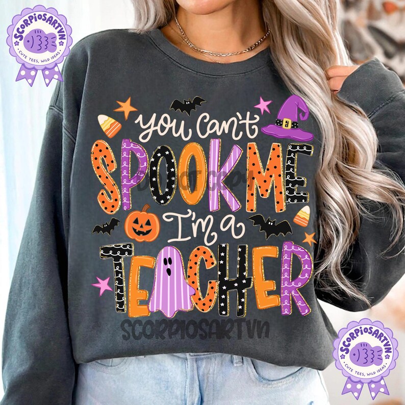 May include: A dark gray crewneck sweatshirt featuring the text "you can't SPOOK ME I'm a TEACHER" in colorful, patterned letters. The design includes Halloween-themed graphics such as a jack-o'-lantern, a purple witch hat, bats, and candy corn.