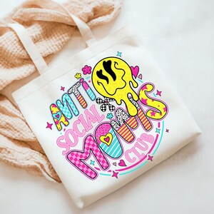 May include: White canvas tote bag with the words "ANTI SOCIAL MOMS CLUB" in a colorful, retro font. The design includes a yellow melting smiley face, hearts, stars, and floral accents. The bag has two white handles.