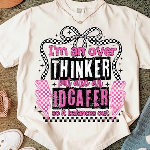 Puede incluir: Camiseta color crema con el texto "I'm an over thinker but also an IDGAF-er so it balances out" en negro y rosa. El dise&ntilde;o incluye un lazo a cuadros, rayos y estrellas.