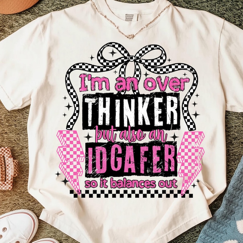 Puede incluir: Camiseta color crema con el texto "I'm an over thinker but also an IDGAF-er so it balances out" en negro y rosa. El dise&ntilde;o incluye un lazo a cuadros, rayos y estrellas.