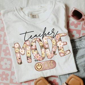 May include: White t-shirt with the text "Teacher Mode Off" in black and pink lettering. The letters are decorated with a floral pattern. A pink and gold power button is below the text.