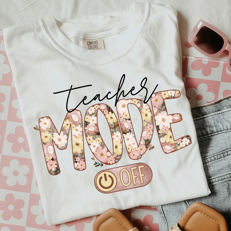 May include: White t-shirt with the text "Teacher Mode Off" in black and pink lettering. The letters are decorated with a floral pattern. A pink and gold power button is below the text.