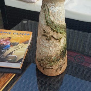 May include: A tall, ceramic vase with a textured, off-white surface and green accents. The vase features a raised design of a dragon wrapping around the body.