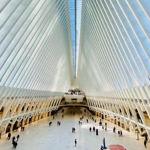 May include: A white, modern, geometric structure with a glass ceiling and a large open space. People are walking around in the space.