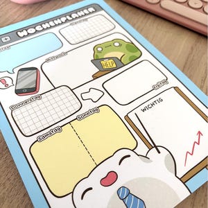 May include: A light blue weekly planner sheet titled Wochenplaner with sections for days of the week. It features cartoon illustrations of a green frog with a laptop labelled HELP and a white character with a blue tie pointing at a Wichtig note.