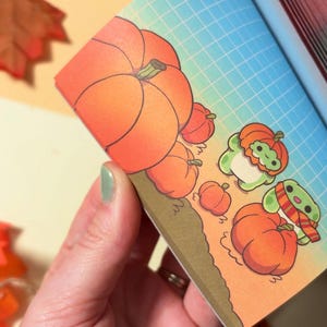 May include: A notepad page featuring a grid background with a gradient from blue to orange. The illustration shows several orange pumpkins and two small green creatures, one wearing a pumpkin hat and the other wearing a striped scarf.