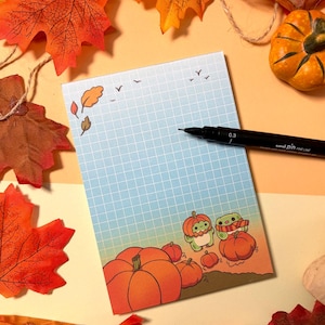 May include: A rectangular notepad with a blue grid pattern, featuring a gradient background and illustrations of small green creatures with pumpkins. A black Uni Pin 0.3 fine line pen rests on the paper, which is surrounded by artificial autumn leaves.
