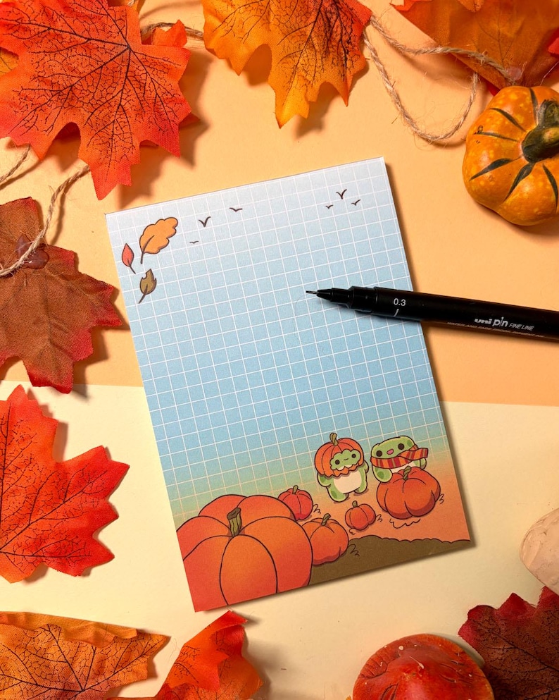 May include: A rectangular notepad with a blue grid pattern, featuring a gradient background and illustrations of small green creatures with pumpkins. A black Uni Pin 0.3 fine line pen rests on the paper, which is surrounded by artificial autumn leaves.