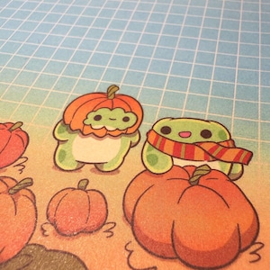 May include: A printed illustration featuring two small green frog-like creatures in a pumpkin patch. One frog wears a pumpkin as a hat, while the other wears a striped scarf and sits atop a large orange pumpkin against a blue grid background.