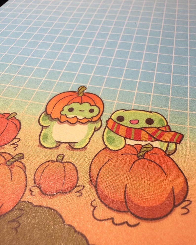 May include: A printed illustration featuring two small green frog-like creatures in a pumpkin patch. One frog wears a pumpkin as a hat, while the other wears a striped scarf and sits atop a large orange pumpkin against a blue grid background.
