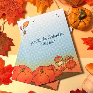 May include: A rectangular notepad featuring a light blue grid pattern, autumn leaves, and pumpkins. The text "gem&uuml;tliche Gedanken bitte hier" is printed in the center. Two small green creatures are illustrated wearing pumpkin hats.