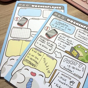 May include: Two light blue weekly planner sheets with a digital interface design. The pages feature sections for each day of the week, including handwritten German text, illustrations of a smartphone, a frog with a help sign, and a smiling creature.