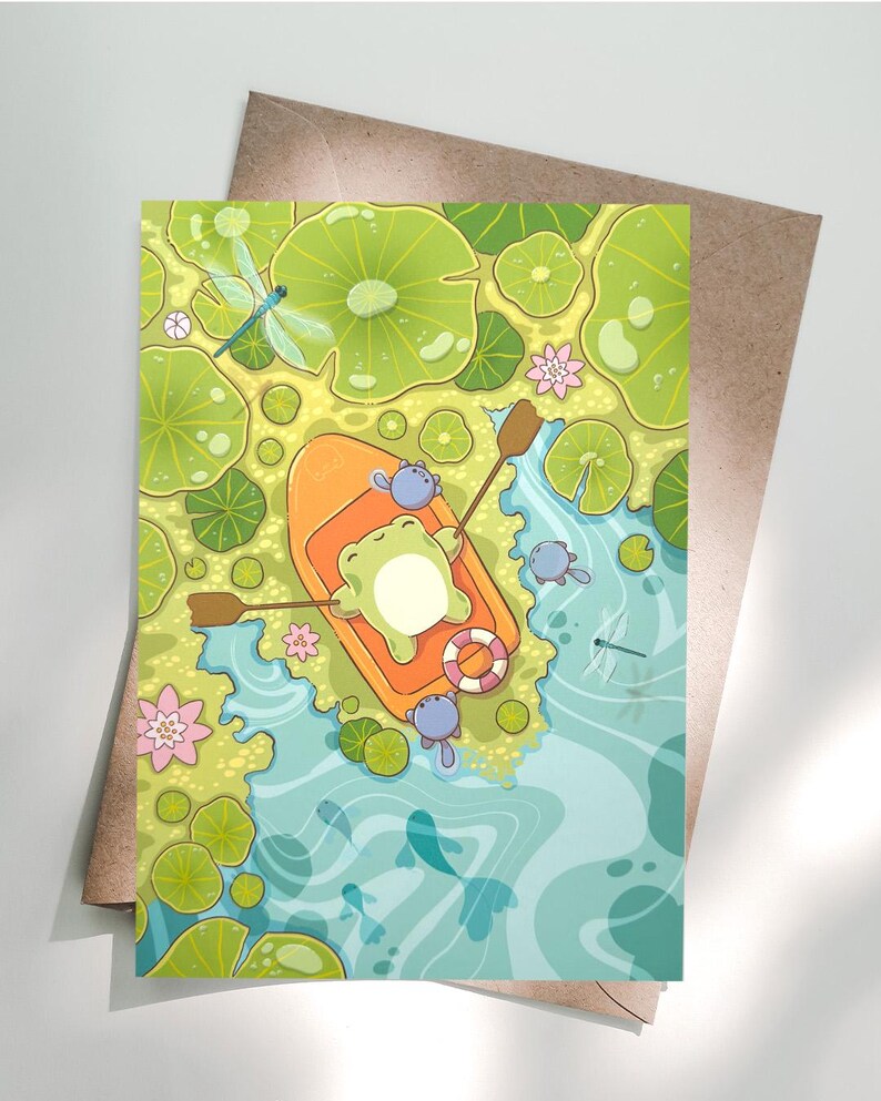 May include: A whimsical illustration of a frog in an orange boat, floating on a pond with lily pads and dragonflies. The frog is wearing a life preserver. The pond has blue water and is surrounded by green lily pads and pink flowers. The illustration is on a greeting card.