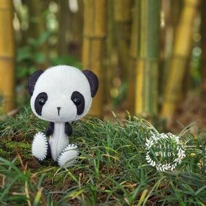May include: A small, white and black felt panda bear sitting in a bed of green grass. The panda has black eyes and a black nose. The background is a blurred image of bamboo.