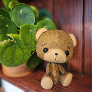 May include: A brown felt teddy bear with black eyes and a stitched brown nose. The bear is sitting on a wooden surface with a green plant in the background.