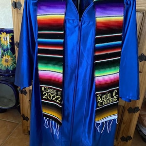 May include: A blue graduation gown with a black and colorful graduation stole. The stole has the text "Class of 2022" and "Kevin C. Thomas" embroidered on it.