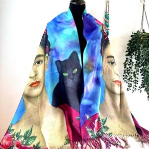 Könnte beinhalten: Ein farbenfroher Schal mit einem Porträt einer Frau, einer schwarzen Katze und floralen Akzenten. Der Schal hat ein blaues, rosa und beiges Farbschema mit Fransen. Der Schal wird auf einer schwarzen Schaufensterpuppe präsentiert.